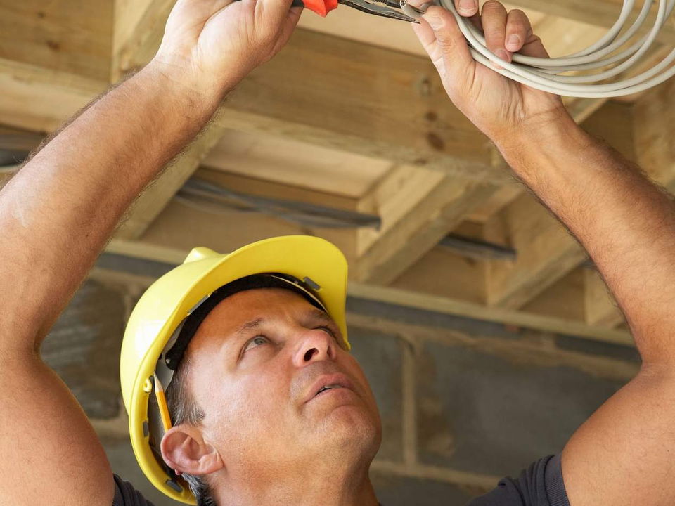 An electrician in a yellow hard hat pulling cable through the ceiling joists of a house