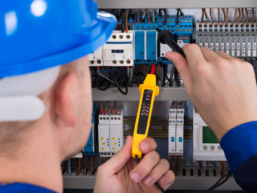 An electrician in a blue hard hat checking a switchboard full of circuit breakers with a yellow tester